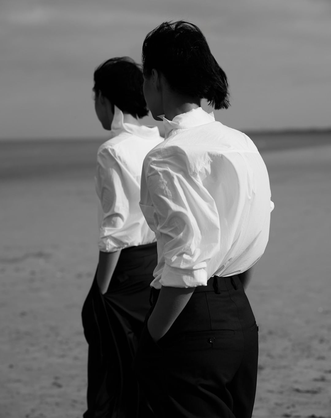 Two people in white shirts and dark trousers standing outdoors on a sandy beach, photographed in black and white by Chris and Cory.