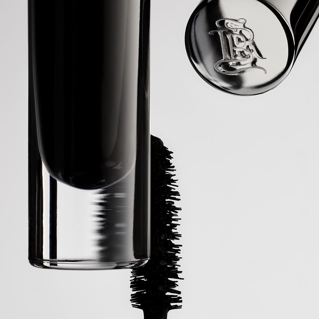 Close-up of a mascara tube and applicator with branding on a white backdrop, showcasing beauty and cosmetic product.