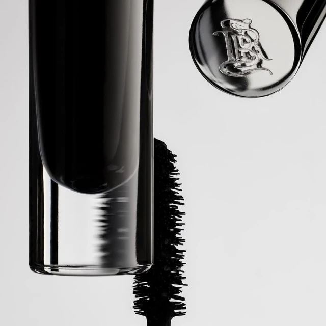 Close-up of a mascara tube and applicator with branding on a white backdrop, showcasing beauty and cosmetic product.