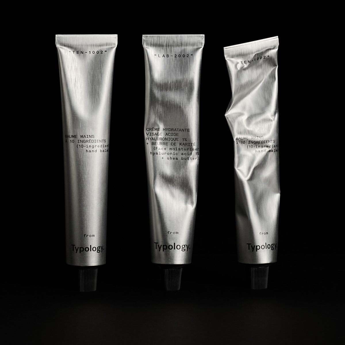 Three silver tubes of Typology hand cream are lined up against a dark background in a product shot showcasing minimalist beauty.