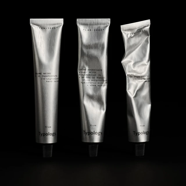 Three silver tubes of Typology hand cream are lined up against a dark background in a product shot showcasing minimalist beauty.