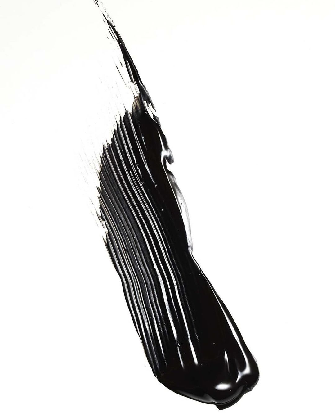Bold stroke of black mascara on white background. The smear has linear striations and glossy texture, creating a minimalist composition.