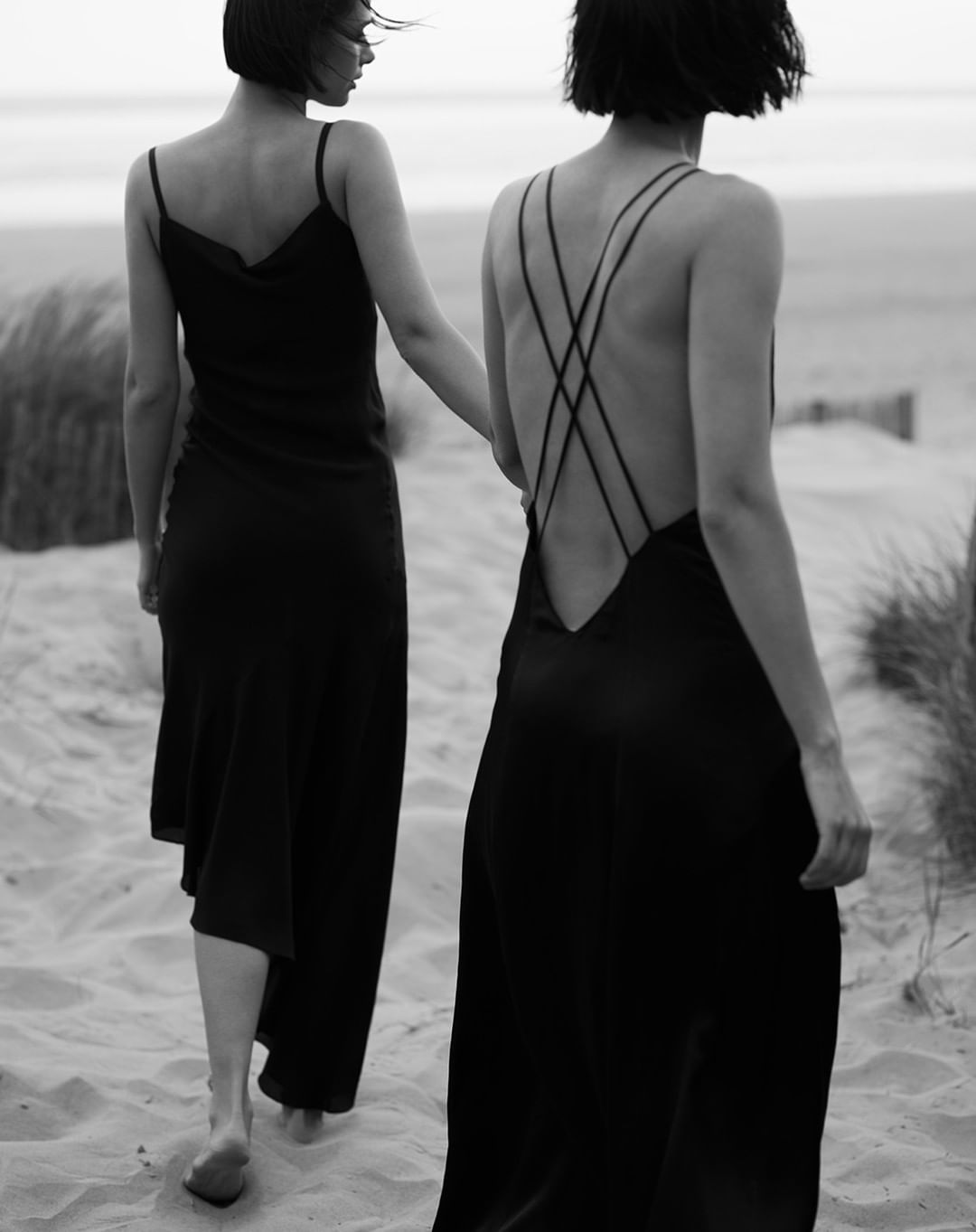 Two women in black dresses walk barefoot on a sandy beach with dune grass in the background, creating a minimalist fashion image.