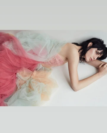 Adult female model lies on a white surface wearing a colorful tulle gown in a studio with soft, neutral lighting for a fashion editorial.