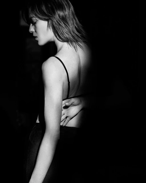 Striking black and white portrait of a woman with short hair, bare back, and hand resting at waist in dark, artistic studio lighting.
