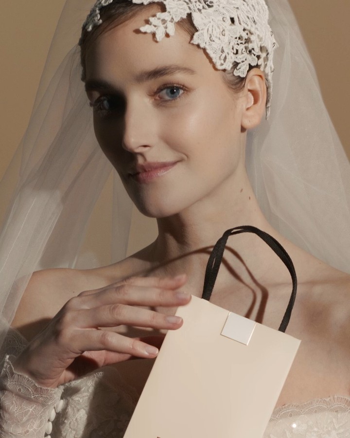 A captivating close-up features a woman in bridal wear, gazing softly while holding a tan gift bag with black handles, set against a warm background.
