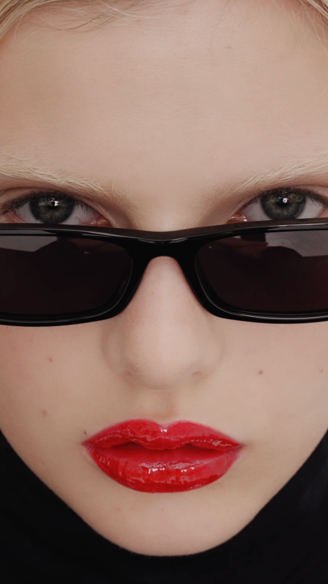 Close-up portrait of a young fair-skinned woman with bright red lips, wearing dark sunglasses and a black turtleneck sweater.