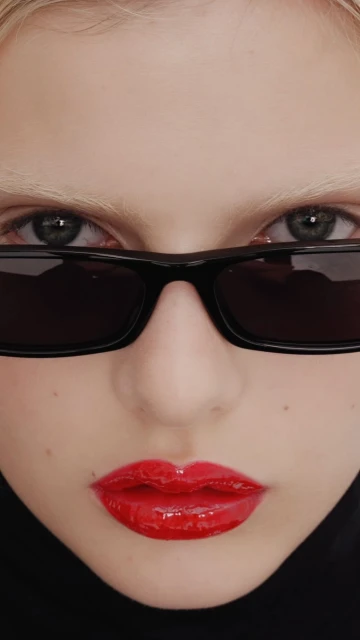 Close-up portrait of a young fair-skinned woman with bright red lips, wearing dark sunglasses and a black turtleneck sweater.