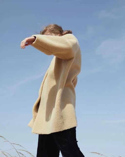 A stylish woman in a tan coat stands in a field of wheat against a blue sky, posing away from the camera in an outdoor setting.