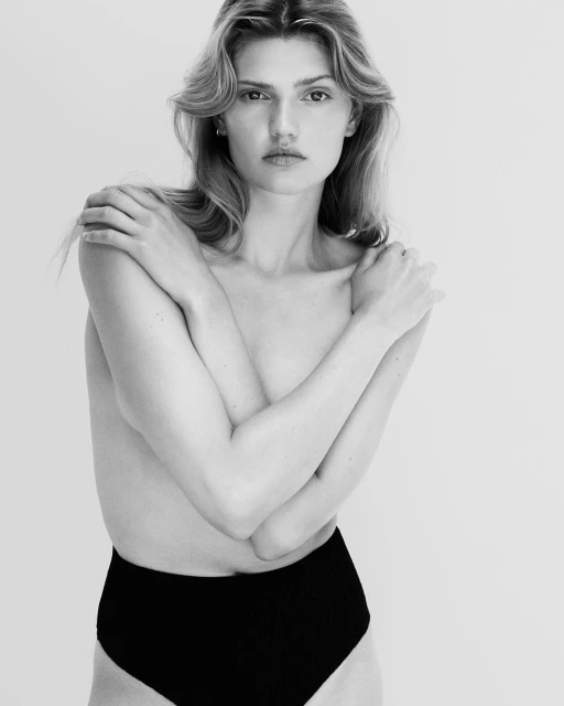 Black and white portrait of a fair-skinned young adult woman with long blonde hair, arms crossed covering her chest, wearing black bottoms.