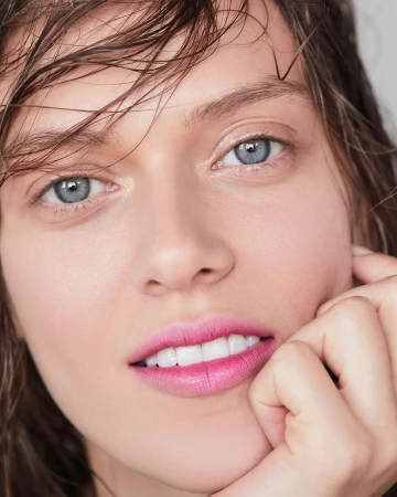 Close-up portrait of a woman with wet hair and pink lipstick resting her hand on her chin against a neutral background