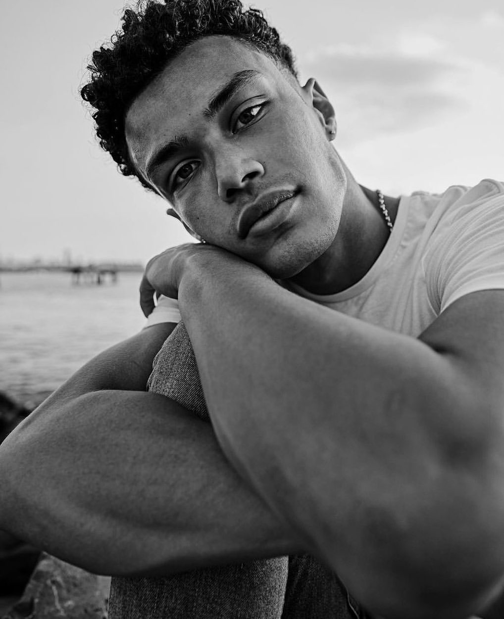 Portrait of a young adult man with curly hair sitting near the water, photographed in black and white with a focus on his face and upper body.