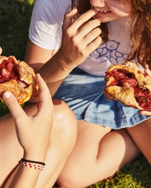 Two people share strawberry galettes on green grass, showcasing a sunlit outdoor scene, light blue denim shorts and casual white t-shirts.