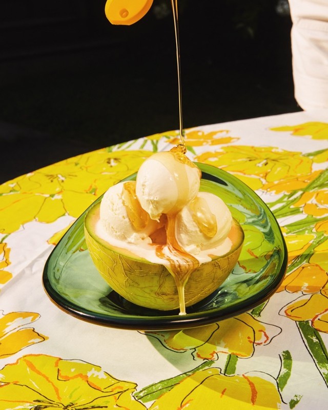 Still life close-up of cantaloupe bowl filled with scoops of vanilla ice cream and topped with honey on a yellow floral tablecloth.