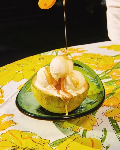 Still life close-up of cantaloupe bowl filled with scoops of vanilla ice cream and topped with honey on a yellow floral tablecloth.
