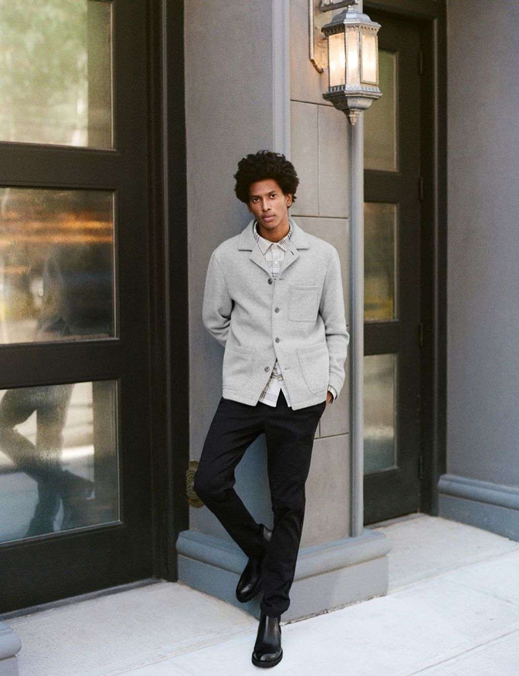 A casually-dressed man with an afro leans against a wall outside next to a doorway. He wears a light jacket, collared shirt, and dark pants.