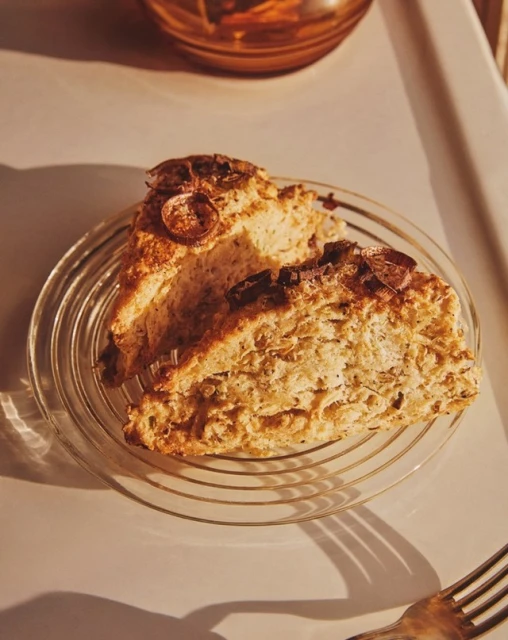 Two pieces of brown scones on a clear plate, next to a silver fork with a bowl in the background, in a studio setting.