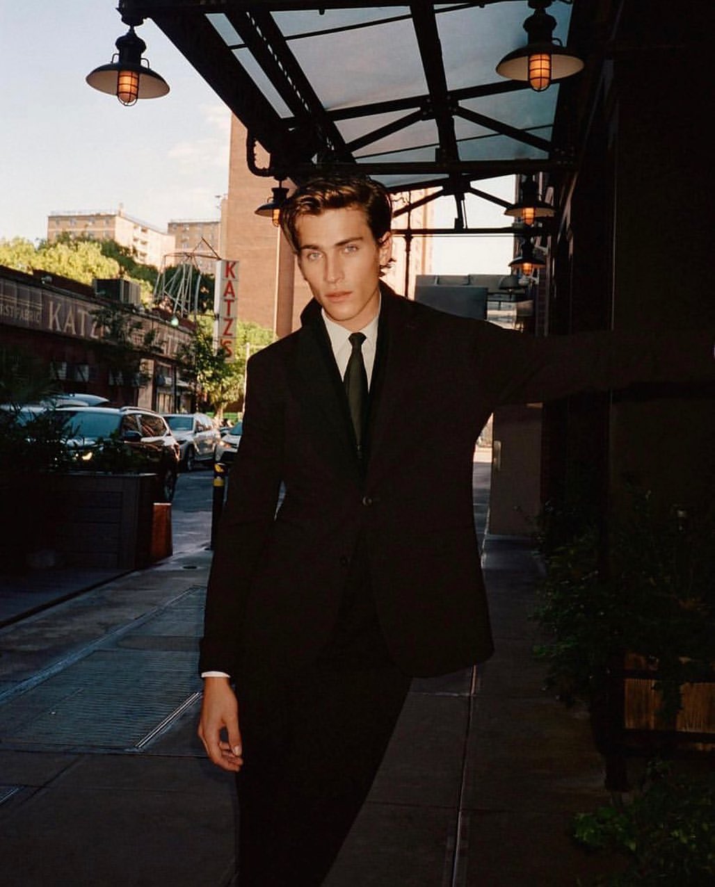 Portrait of a young adult male in a dark suit with a tie, posing outdoors on a city street with buildings and vehicles in the background.