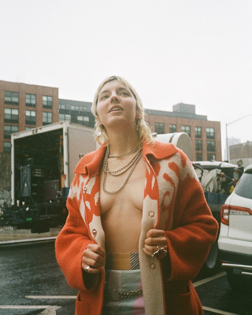 Woman in an orange patterned coat poses outdoors in a low-angle shot with urban background, wearing multiple silver necklaces and rings.