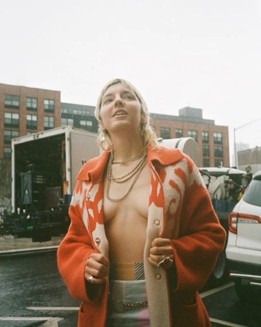 Woman in an orange patterned coat poses outdoors in a low-angle shot with urban background, wearing multiple silver necklaces and rings.