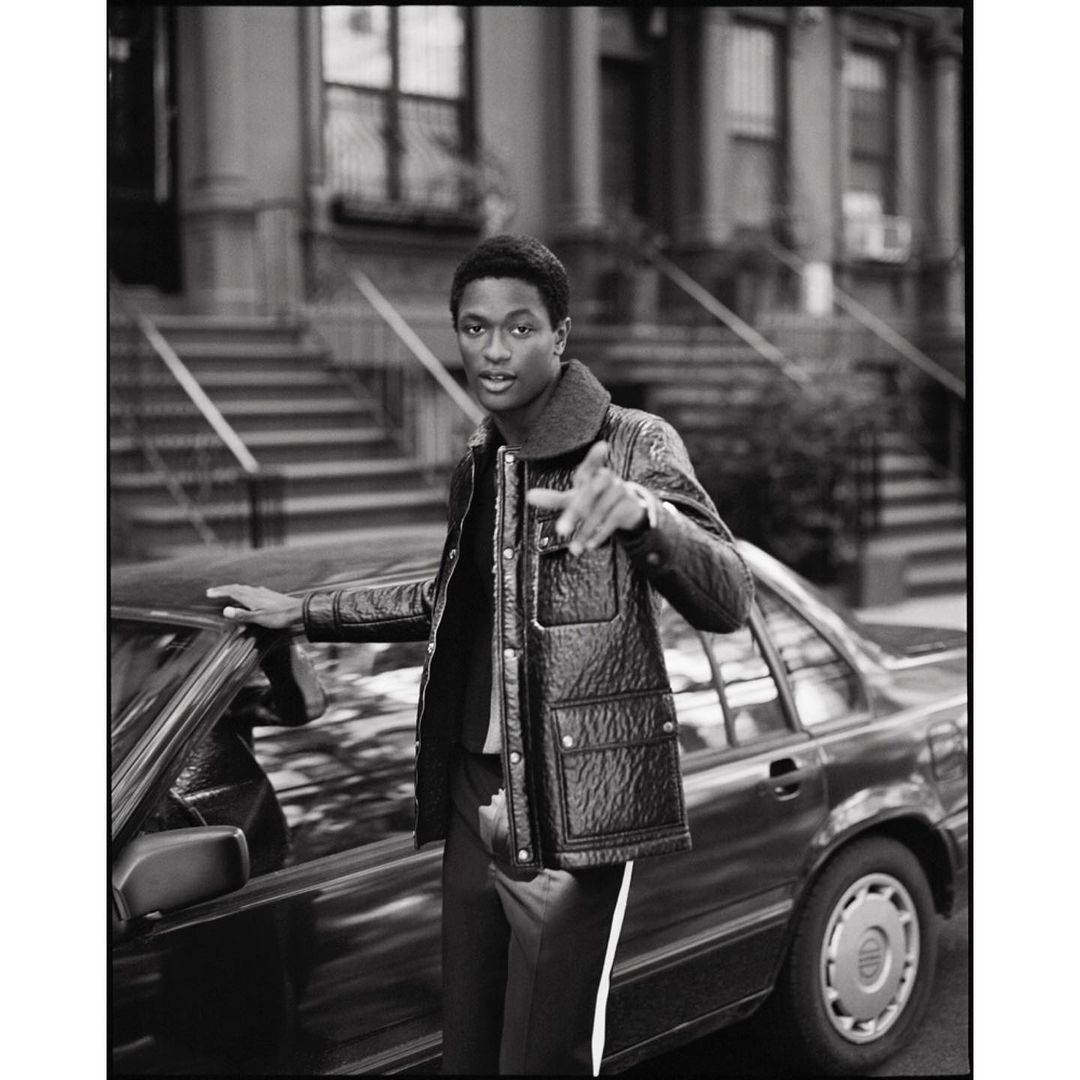 Monochrome shot of a young Black man in a jacket pointing towards the camera near a car and a residential building with steps.