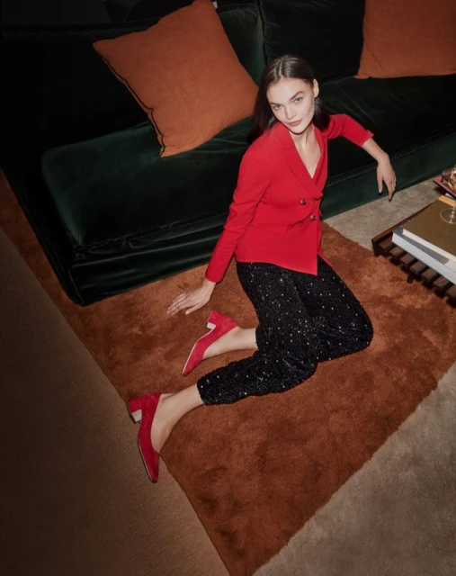 Woman in red jacket and black sequin pants sits on a brown rug near a green velvet sofa, creating a stylish fashion scene.