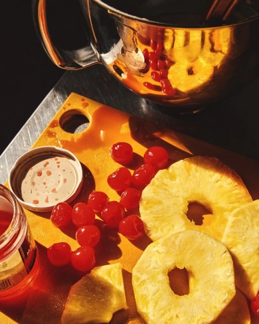 Overhead shot of a wooden cutting board with pineapple slices, maraschino cherries, dipping sauce and chrome serving dish.