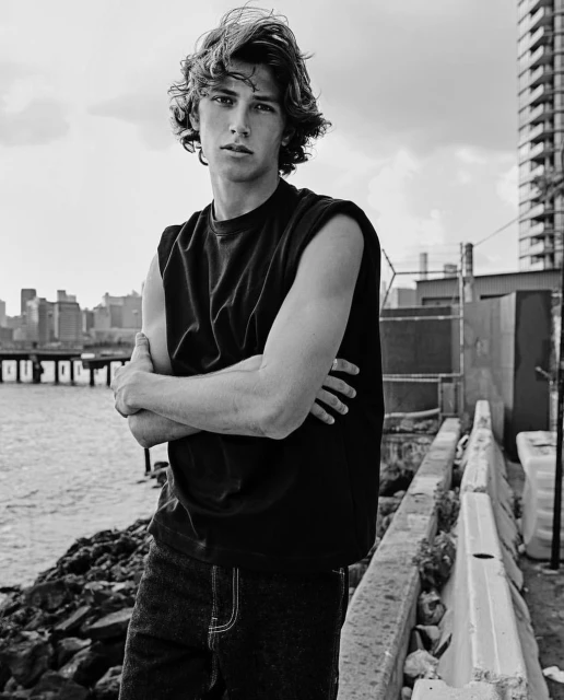 Portrait of a young adult man in a sleeveless shirt with arms crossed. The background includes a cityscape and waterfront.