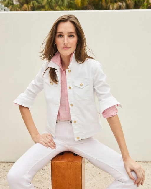 Elegant woman in a white denim jacket and pants poses on a wood box against a white wall with green foliage overhead.