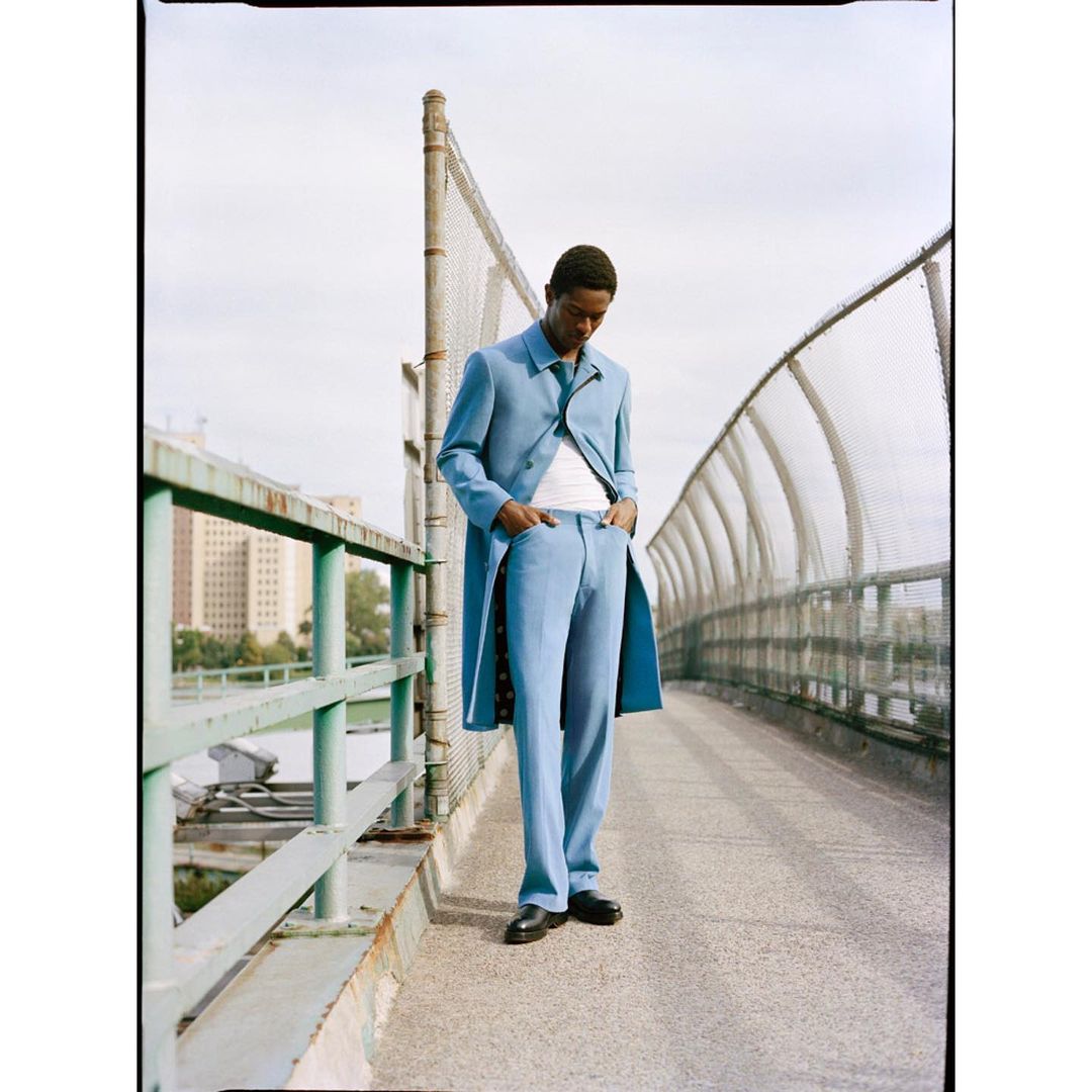 Man poses in a light blue coat and pants on a fenced walkway with buildings and a cloudy sky in the background. Fashion editorial.
