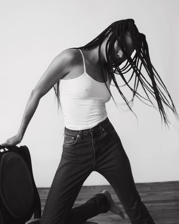 Black and white portrait of a woman with long braided hair whipping through the air while wearing a tank top and jeans.