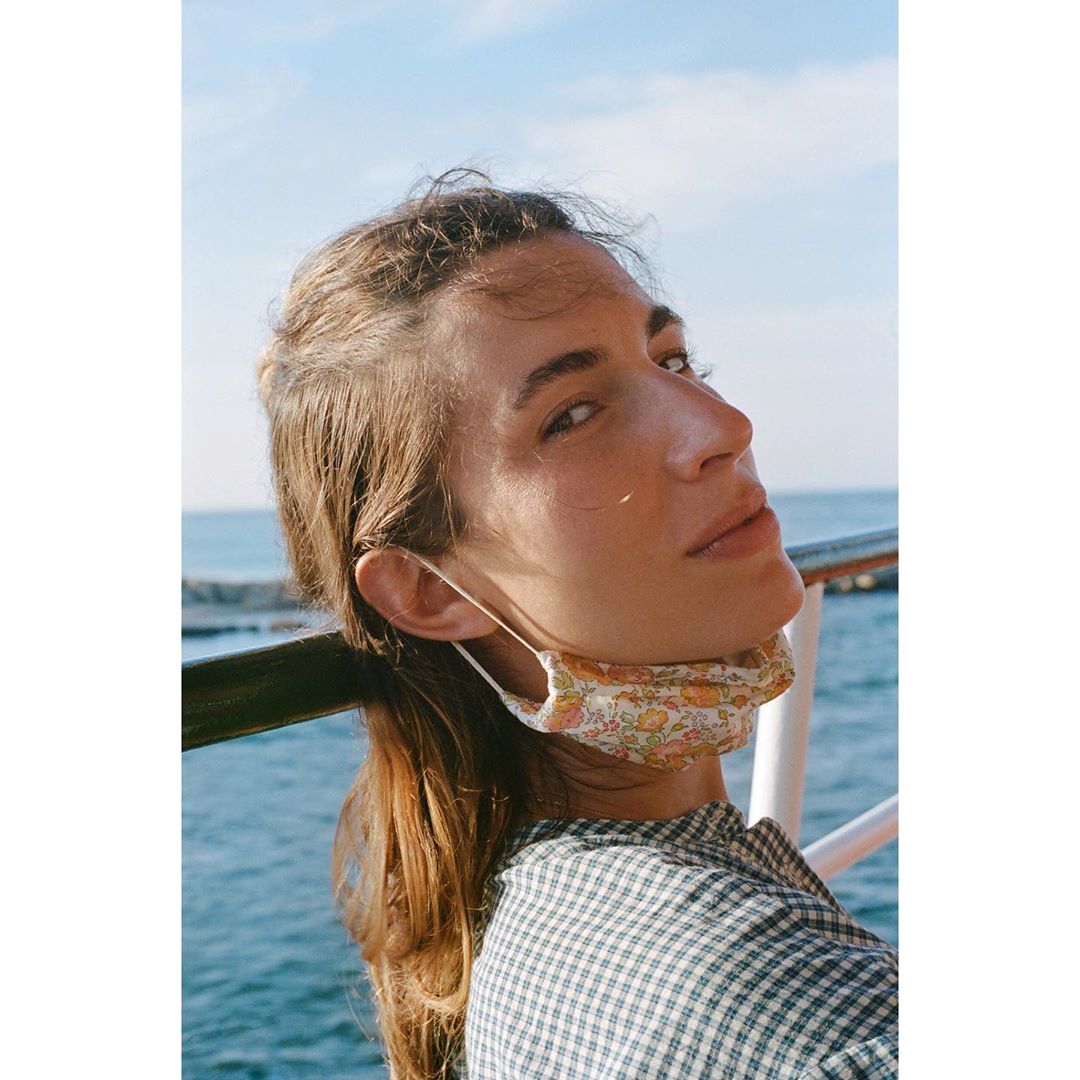 Portrait of an adult woman wearing a floral face mask while standing outdoors, featuring the ocean and blue sky in the background.