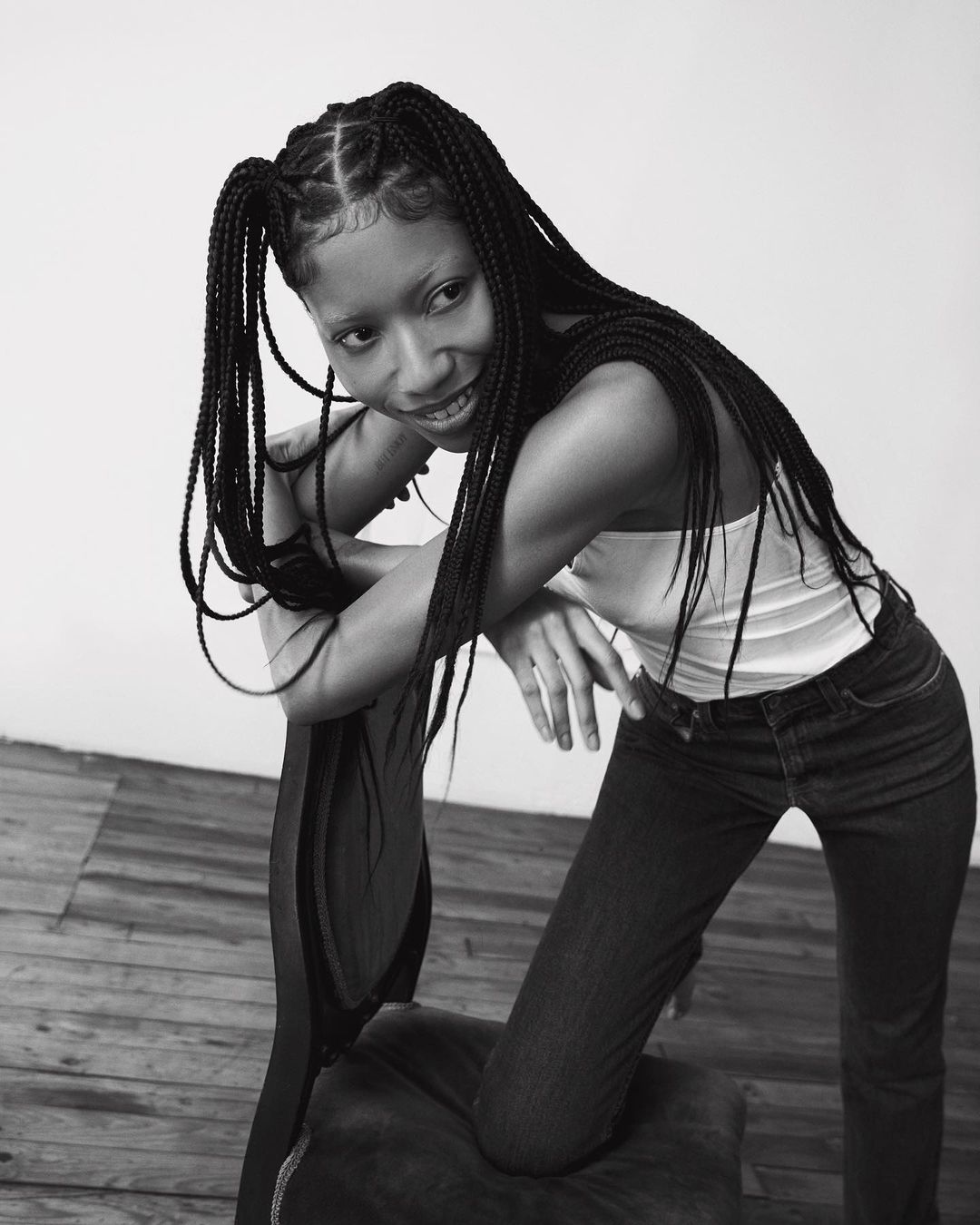 Black and white portrait of a woman with braided hair leaning over a chair, wearing a white tank top and dark jeans.