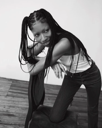 Black and white portrait of a woman with braided hair leaning over a chair, wearing a white tank top and dark jeans.