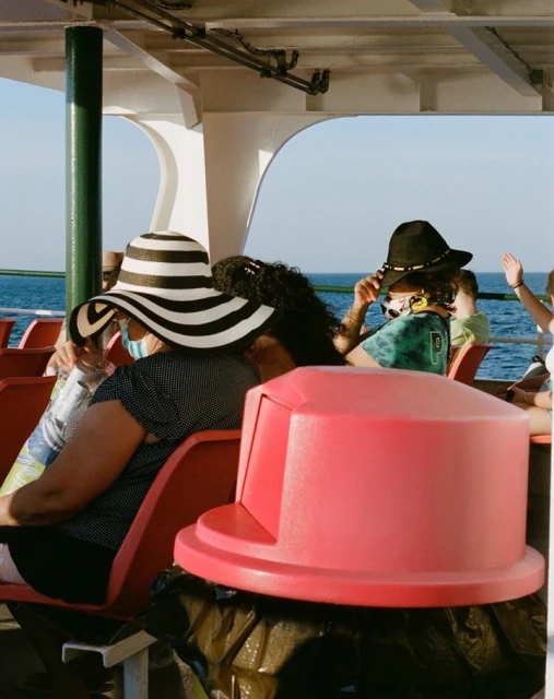 Passengers on a ferry include two people wearing hats in the foreground, with a pink trash can at the front of the frame.