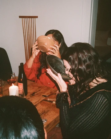 Two women drinking from bowls indoors at a candlelit table with wine and tableware, enjoying a casual meal together.