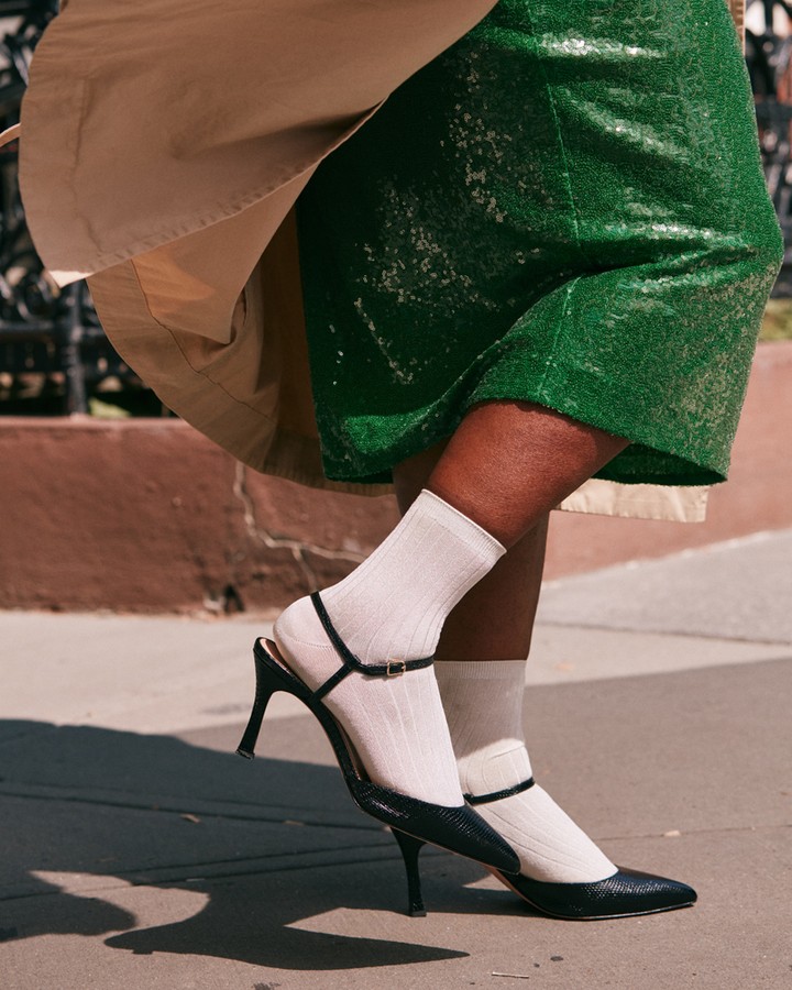 Woman showcases green sequin skirt, trench coat, white ribbed socks, and black high-heeled T-strap pumps on a city street.