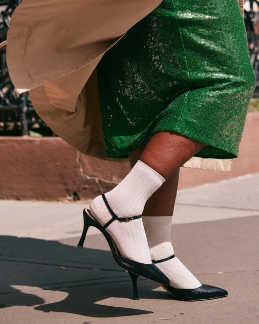 Woman showcases green sequin skirt, trench coat, white ribbed socks, and black high-heeled T-strap pumps on a city street.