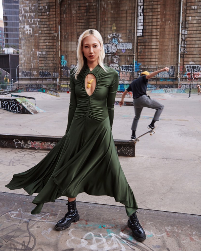 Young woman in long green dress and black boots poses at skate park with skateboarder in background; urban fashion editorial photography.