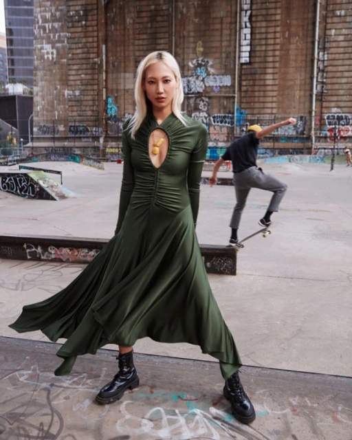 Young woman in long green dress and black boots poses at skate park with skateboarder in background; urban fashion editorial photography.