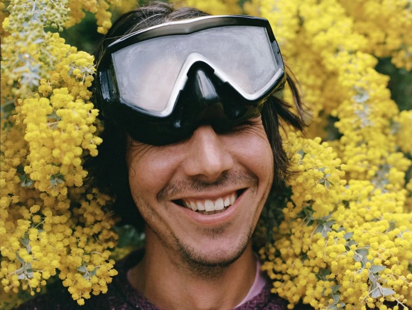 Smiling adult male with dark hair and ski goggles surrounded by vibrant yellow flowers in an outdoor environment, close-up shot.