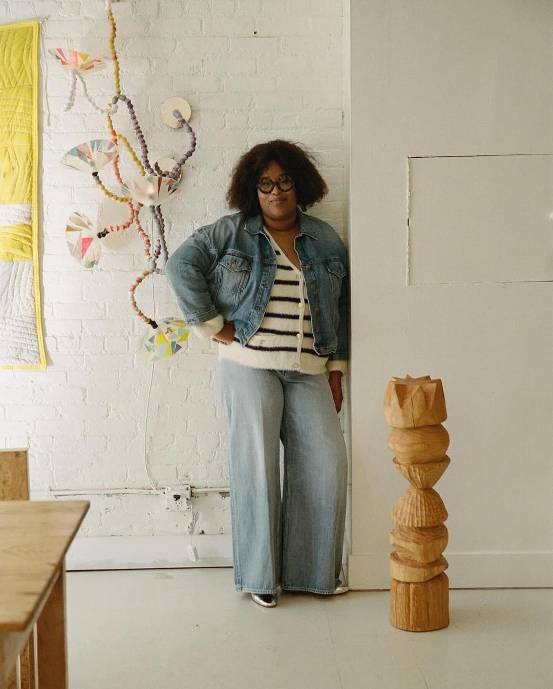 A woman in a denim jacket and wide-leg jeans poses indoors near a wooden sculpture and wall art, looking directly at the camera.