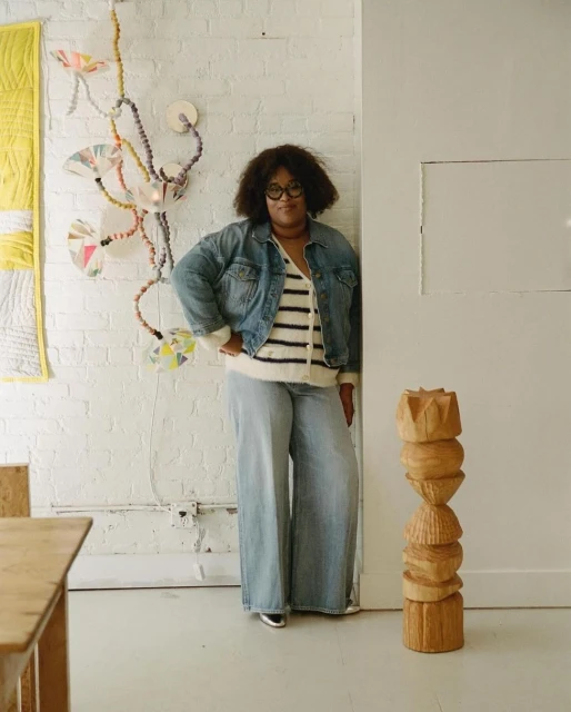 A woman in a denim jacket and wide-leg jeans poses indoors near a wooden sculpture and wall art, looking directly at the camera.