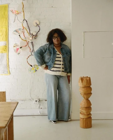A woman in a denim jacket and wide-leg jeans poses indoors near a wooden sculpture and wall art, looking directly at the camera.