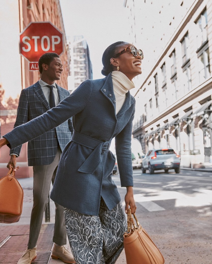 Stylish African American woman in a blue coat laughs while walking on a city street with a man carrying a bag and a blurred car.