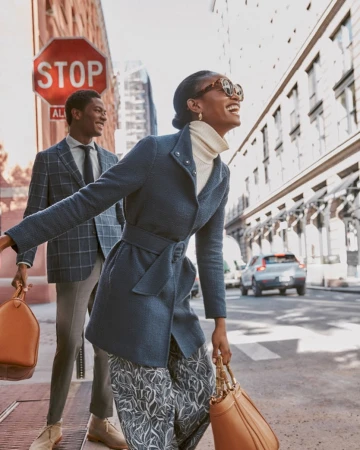 Stylish African American woman in a blue coat laughs while walking on a city street with a man carrying a bag and a blurred car.