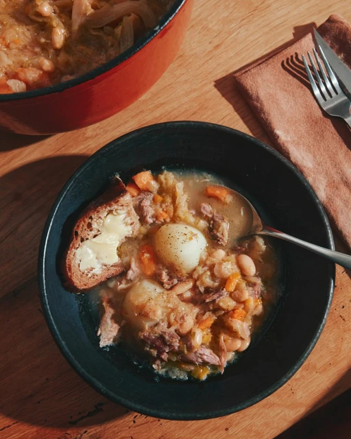 Overhead shot of hearty stew with potatoes, beans, and meat in a black bowl with bread on a wooden table next to silverware.