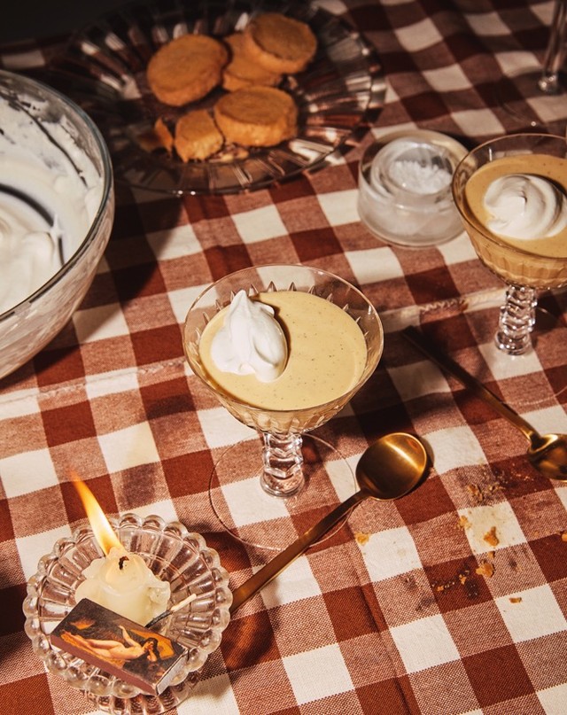 Overhead shot of dessert glasses filled with custard and whipped cream on a red-and-white checkered tablecloth, with cookies and candle.