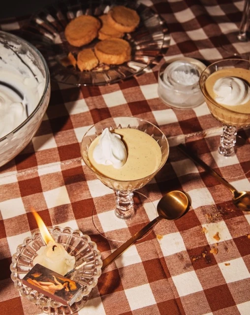 Overhead shot of dessert glasses filled with custard and whipped cream on a red-and-white checkered tablecloth, with cookies and candle.
