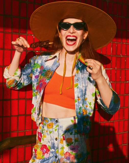 Woman in a floral outfit and oversized hat eating meat on a stick against a red tiled wall, sunglasses on, smiling broadly.